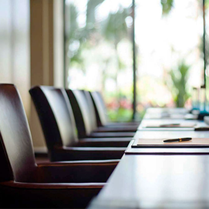 A row of empty chairs at a conference table in a brightly lit room with large windows and an outdoor view.