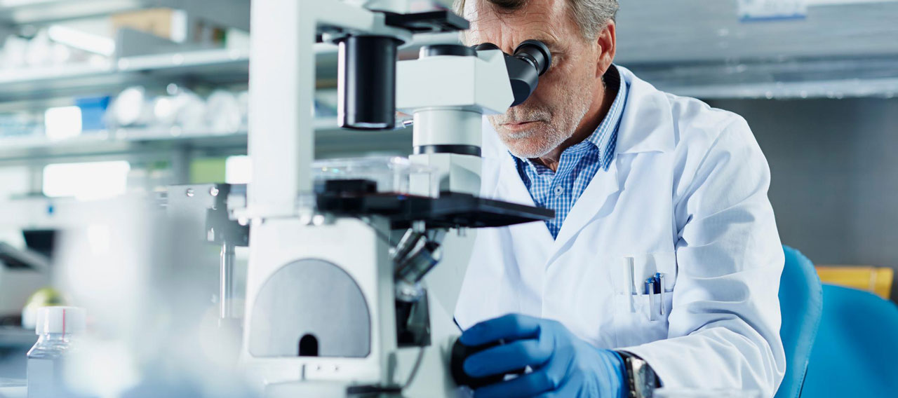 A man in a lab coat examines a sample through a microscope in a laboratory setting.